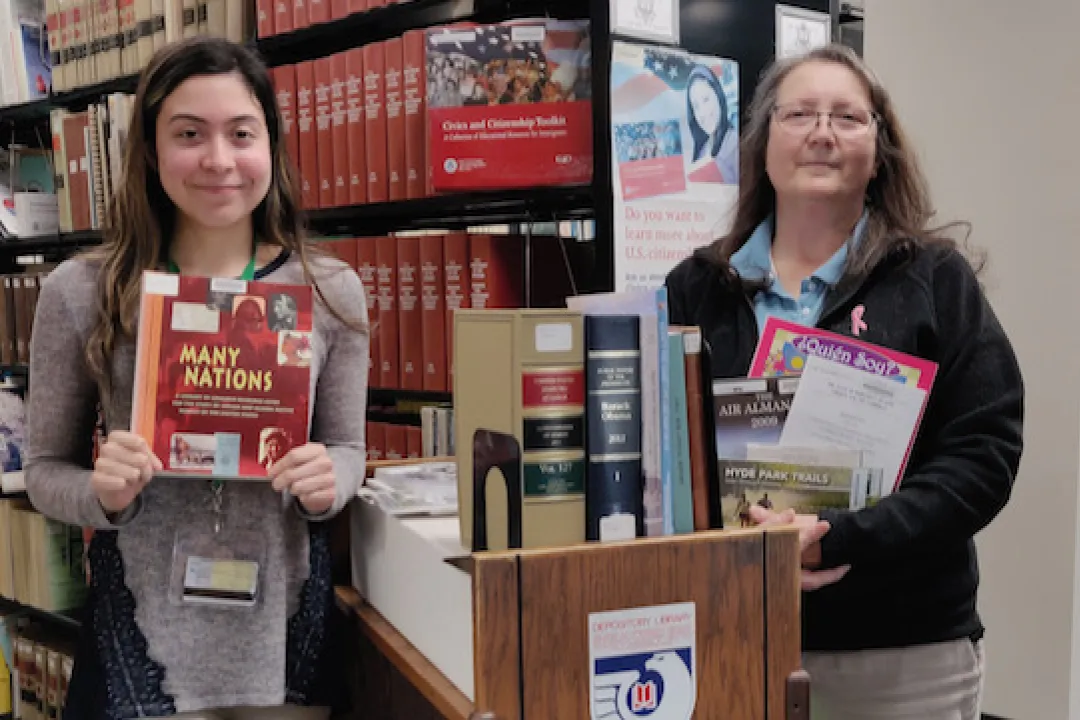 Two women in front of book shelves holding up government publications.